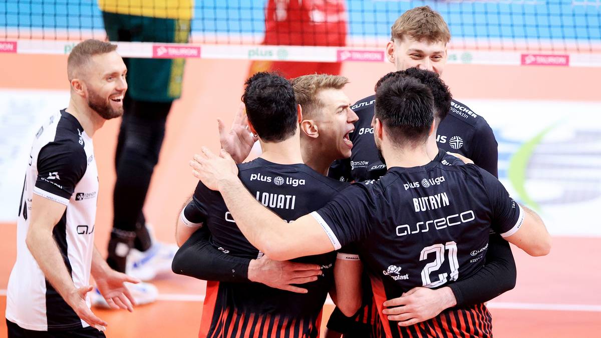 Sześciu siatkarzy w czarnych strojach z czerwonymi pasami, świętujących przy siatce.