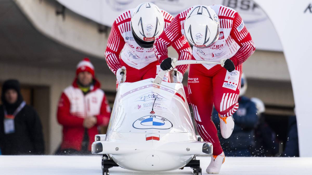 Dwóch zawodników w kombinezonach pcha sanie bobslejowe