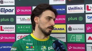 Portret mężczyzny w zielonej koszulce sportowej z ż&oacute;łtym kołnierzem, patrzącego w bok. W tle widoczne są panele z logotypami sponsor&oacute;w.