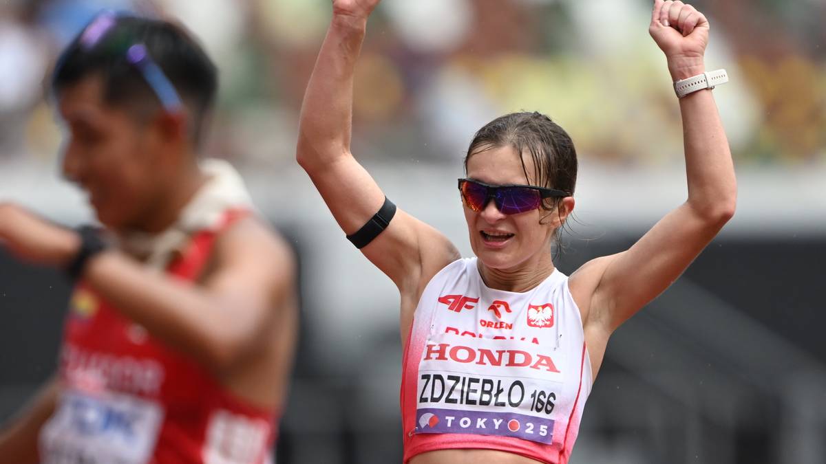 Kobieta w stroju sportowym z rękami uniesionymi w geście radości, na jej koszulce widnieje napis "HONDA ZDZIEBŁO 166 TOKYO 2020". W tle widać zamazaną sylwetkę innego sportowca.