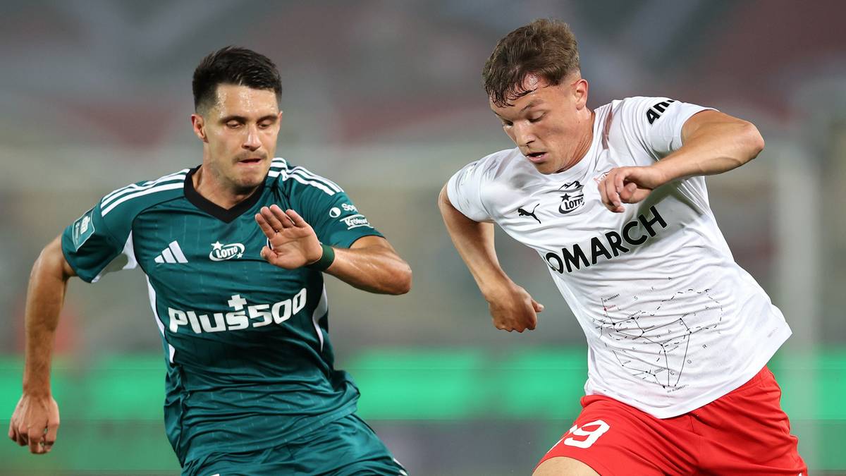 Dwóch piłkarzy w akcji na boisku. Po lewej stronie zawodnik w zielonej koszulce z białymi pasami na rękawach i napisem "PLUS500", po prawej zawodnik w białej koszulce z napisem "OMARCH". Obaj zawodnicy wydają się skupieni i walczą o piłkę.