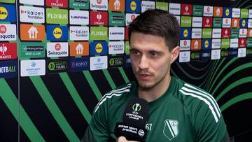 Bartosz Kapustka, piłkarz, odpowiada na pytanie trzymając mikrofon przed ścianą z logotypami sponsor&oacute;w.