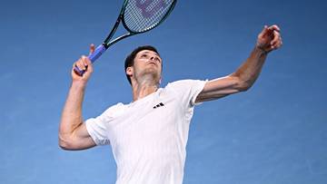 Tenisista w białej koszulce wykonuje podanie z rakietą tenisową nad głową.