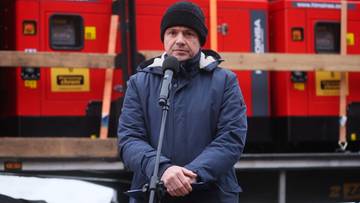 Mężczyzna w czapce i kurtce stoi przed mikrofonem, za nim widać rząd czerwonych agregat&oacute;w prądotw&oacute;rczych.