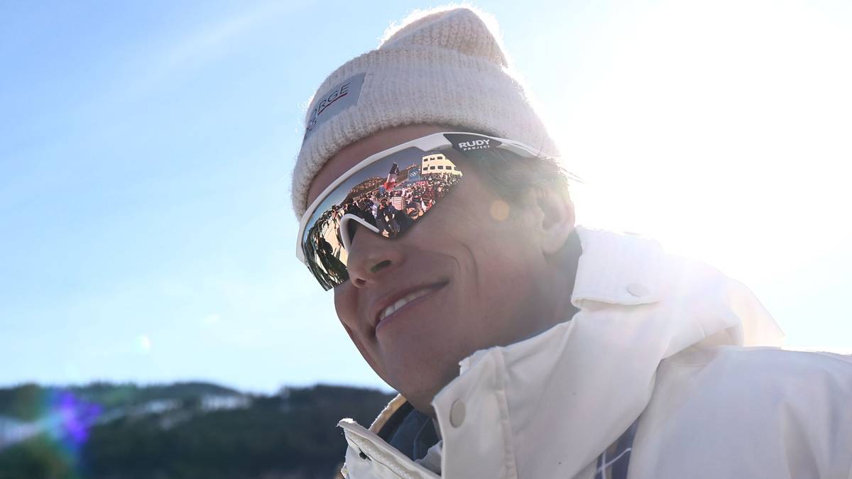 Narciarz biegowy w czapce i goglach narciarskich, z odbiciem stadionu w soczewkach okular&oacute;w.