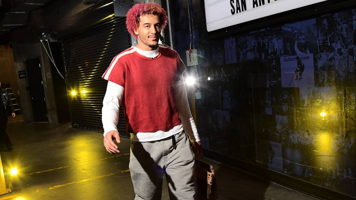 Jeremy Sochan w czerwono-białej bluzie i szarych spodniach idzie korytarzem, na kt&oacute;rym wisi tablica z napisem "crypto.com LOS ANGELES LAKERS VS SAN ANTONIO SPURS".