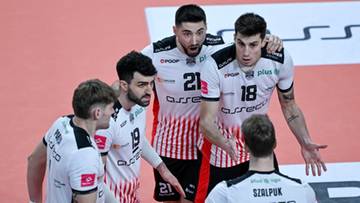 Pięciu siatkarzy w białych koszulkach z czerwonymi pasami, rozmawiających ze sobą na boisku siatkarskim.