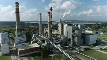 Widok z lotu ptaka na kompleks elektrowni z wieloma kominami, z kt&oacute;rych jeden emituje dym. Widoczne budynki przemysłowe i sieć rurociąg&oacute;w.