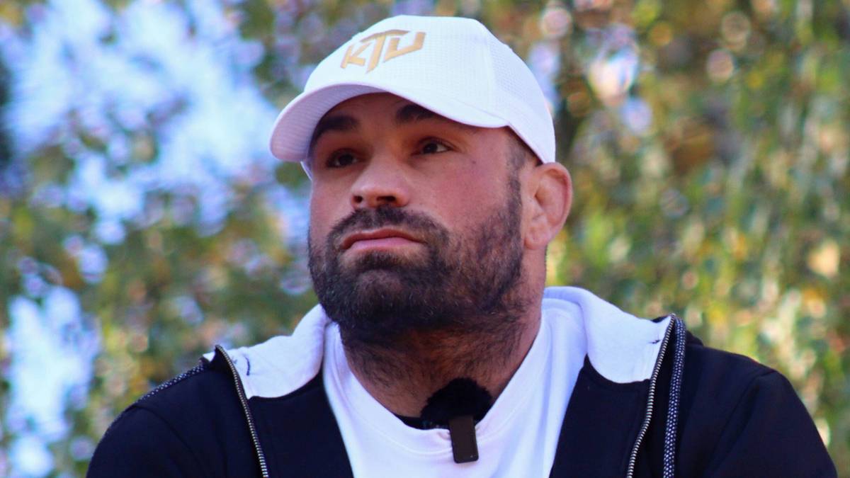 Mężczyzna w czapce i bluzie z kapturem, oparty o barierkę, ze splecionymi dłońmi.