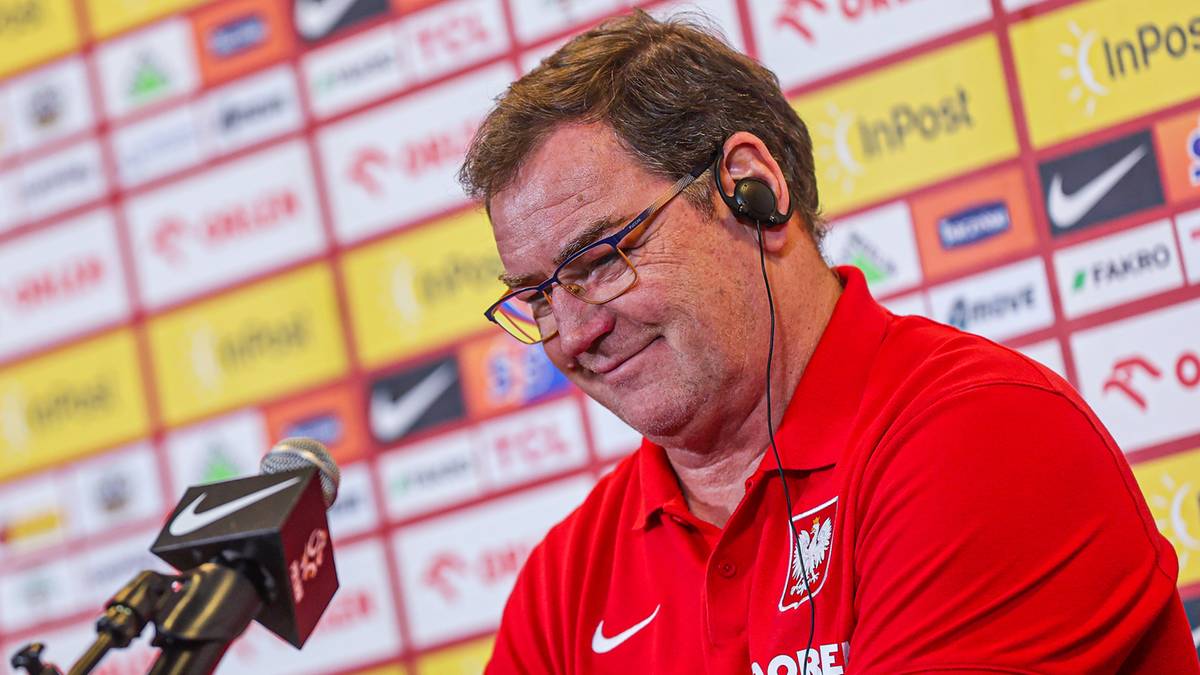 Michał Probierz w czerwonej koszulce reprezentacji Polski, siedzący przy stole z mikrofonem, z okularami i słuchawkami na uszach.