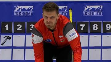 Curler w czerwonej kurtce sportowej kuca na lodowisku, przygotowując się do rzutu kamieniem.