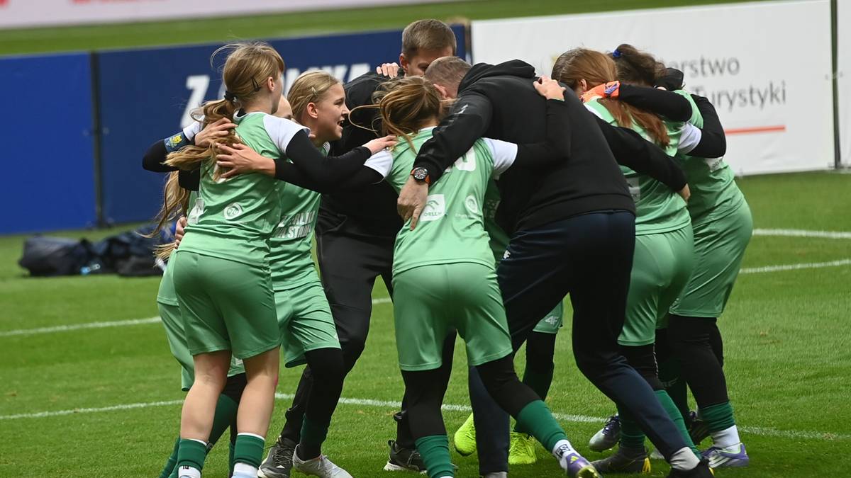 Dziewczęca drużyna piłkarska w zielonych strojach tworzy radosny uścisk na boisku.