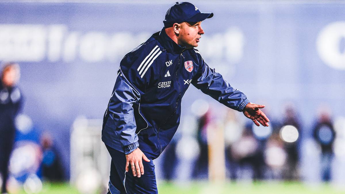 Trener piłkarski w stroju sportowym, zwracający się gestem ręki w bok, na tle murawy i nieostrych sylwetek ludzi.