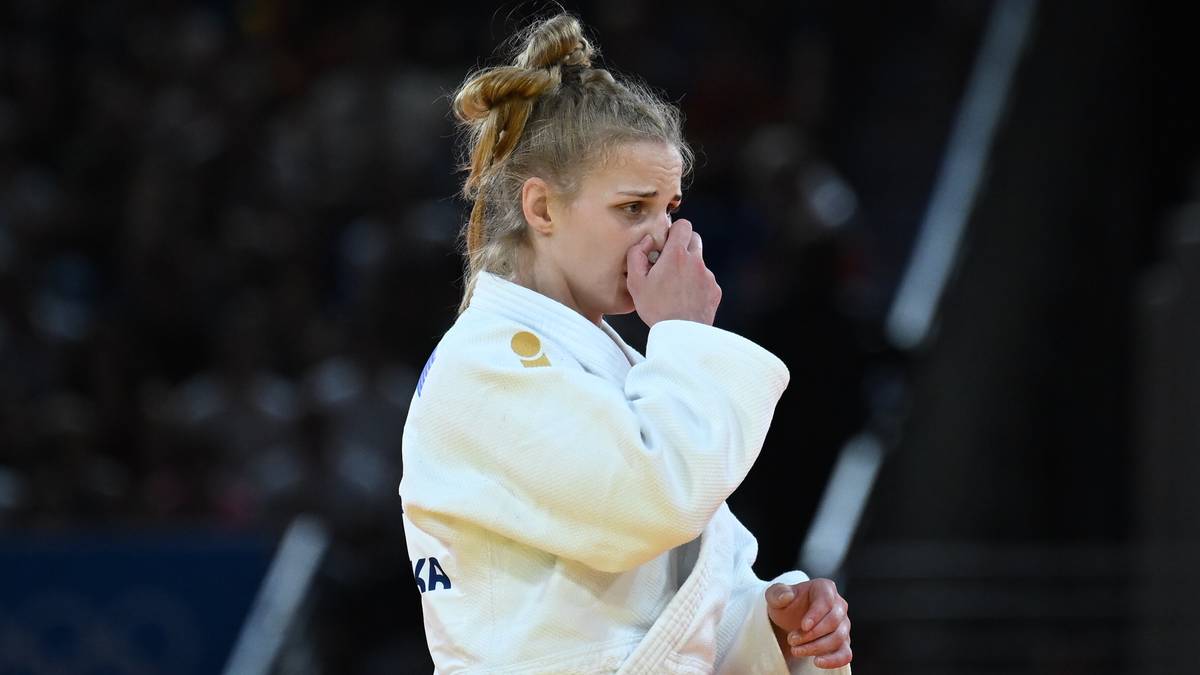 Zawodniczka judo w białym kimonie, z czarnym pasem, zakrywająca usta dłonią, z włosami upiętymi w kok.