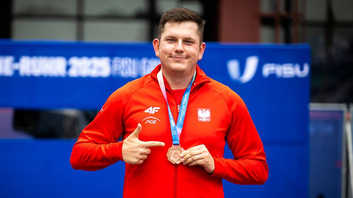 Mężczyzna w czerwonej kurtce sportowej z polskim godłem, uśmiechnięty, trzyma w dłoni medal i wskazuje na niego palcem.