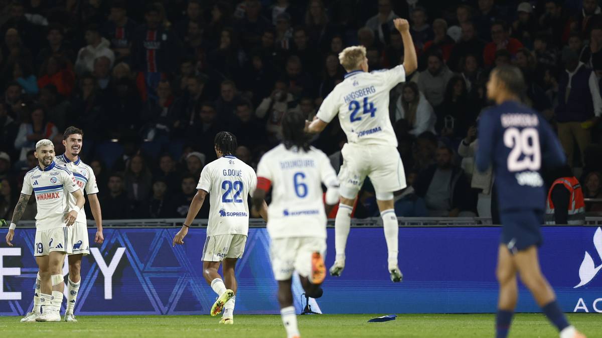 Piłkarze w białych strojach świętują zdobycie bramki podczas meczu piłki nożnej. Jeden z nich skacze z radości, unosząc ręce w górę, podczas gdy inni stoją obok niego. W tle widać trybuny wypełnione kibicami.