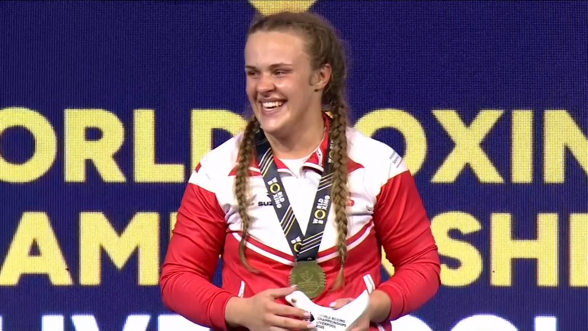 Młoda kobieta w stroju sportowym, z warkoczami, uśmiecha się szeroko, trzymając w dłoniach medal na wstędze i biały przedmiot, na tle napisu "WORLD BOXING CHAMPIONSHIP"