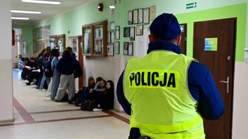Policjant w ż&oacute;łtej kamizelce odblaskowej z napisem POLICJA stoi na korytarzu szkolnym, obserwując grupę uczni&oacute;w siedzących pod ścianą.