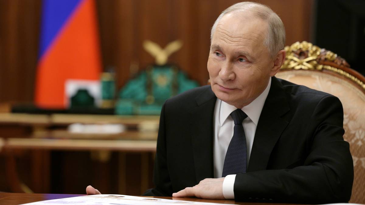 Prezydent Rosji Władimir Putin siedzi przy biurku, ubrany w czarny garnitur i niebieski krawat. W tle widoczna jest flaga Rosji.