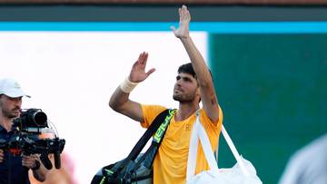 Tenisista Carlos Alcaraz wznosi ręce w geście pozdrowienia, trzymając torbę sportową.