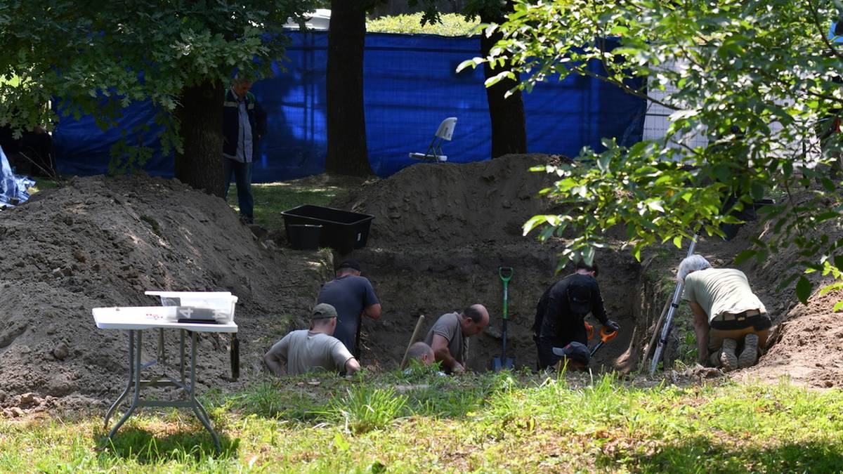 Grupa os&oacute;b prowadzi prace archeologiczne, kopiąc w ziemi. W tle widoczny jest niebieski materiał i drzewa.