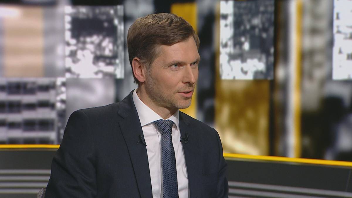 Mężczyzna w garniturze i krawacie siedzi i m&oacute;wi.