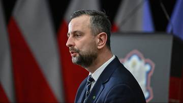 Mężczyzna w garniturze z brodą, stojący na tle flag.