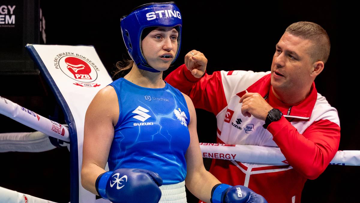 Młoda bokserka w niebieskim stroju i kasku ochronnym, patrząca przed siebie z determinacją. Obok niej stoi trener w czerwono-białej kurtce, z wyrazem skupienia na twarzy, wskazując palcem.