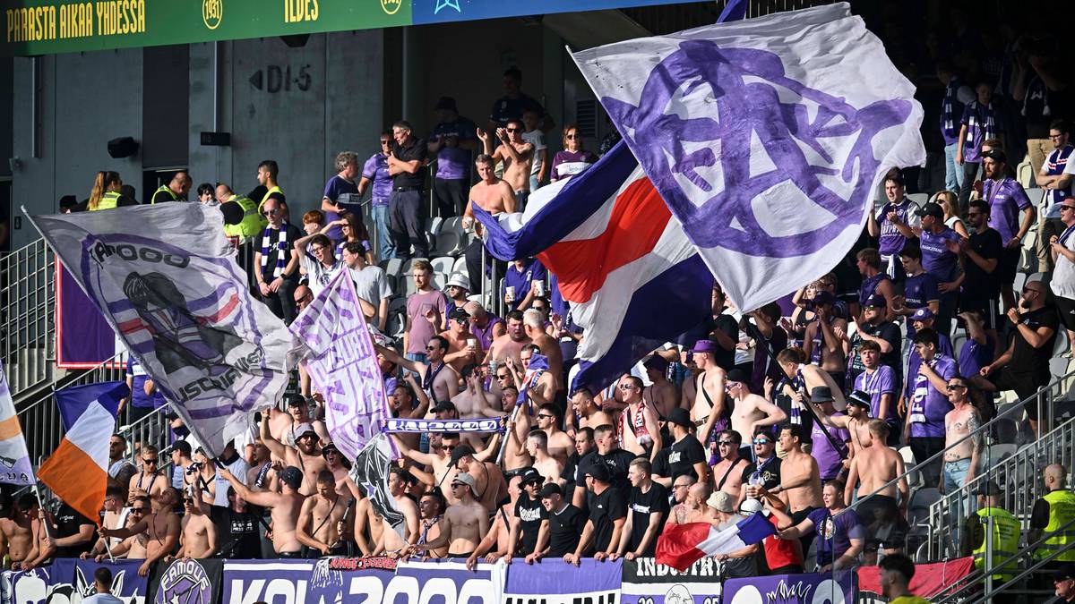 Tłum kibiców na stadionie, trzymających flagi z symbolami drużyny. Jeden z kibiców nosi koszulkę z napisem "Mikołaj Sawicki".