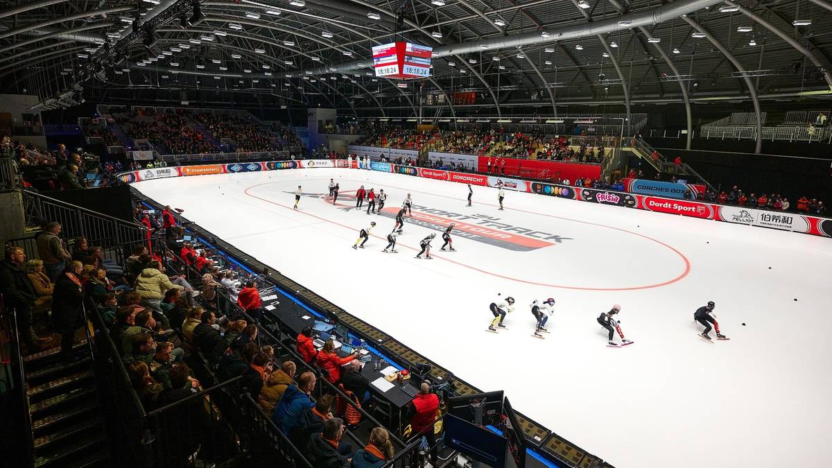 Widok na halę sportową z lodowiskiem do short tracku, na którym odbywają się zawody z udziałem zawodników i widzów na trybunach.