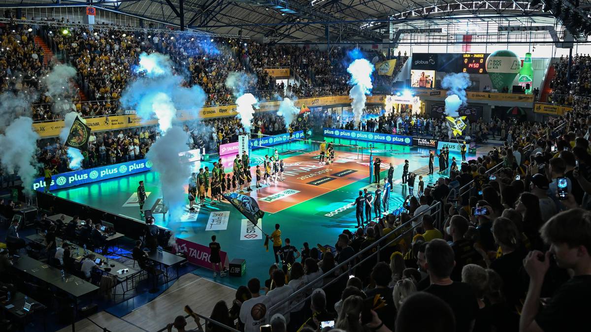 Widok z góry na boisko do siatkówki podczas meczu, otoczone przez pełne trybuny kibiców. Nad boiskiem unoszą się kłęby dymu, tworząc efektowną atmosferę.