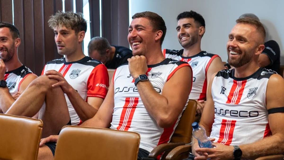 Grupa mężczyzn w biało-czerwonych strojach sportowych siedzi razem, uśmiechając się do kamery. Jeden z nich trzyma w ręku butelkę wody.