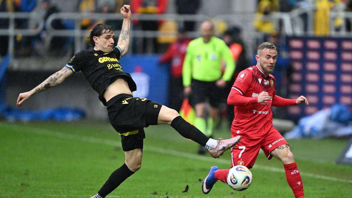 Piłkarz w czarnej koszulce wykonuje wślizg, pr&oacute;bując odebrać piłkę graczowi w czerwonej koszulce.