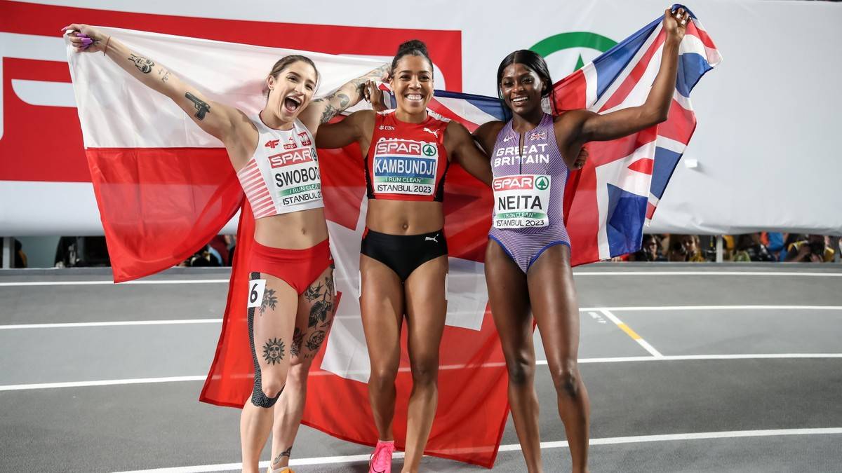 Trzy lekkoatletki pozują do zdjęcia z flagami, świętując zwycięstwo.