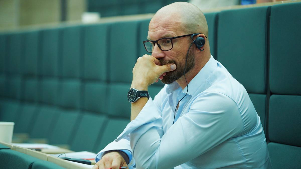 Mężczyzna w okularach i słuchawkach siedzi z ręką na ustach, patrząc w bok, ubrany jest w białą koszulę.