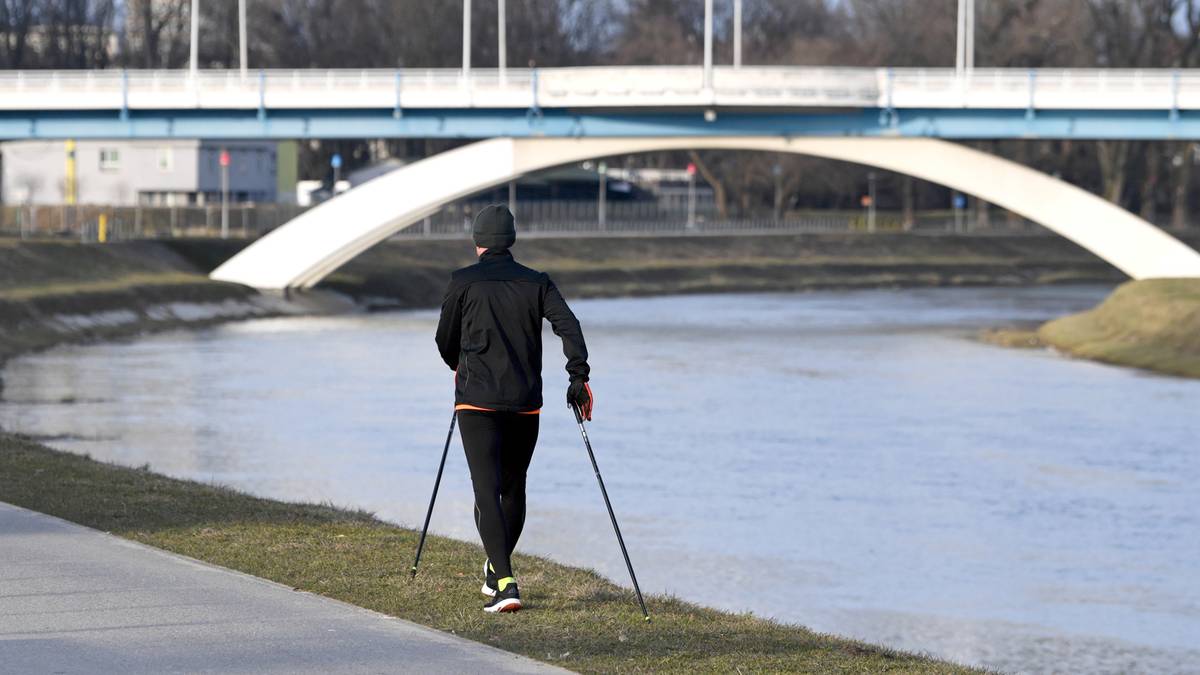 Osoba w czapce i ciemnym ubraniu spaceruje z kijkami do nordic walking wzdłuż rzeki, pod białym łukowatym mostem.