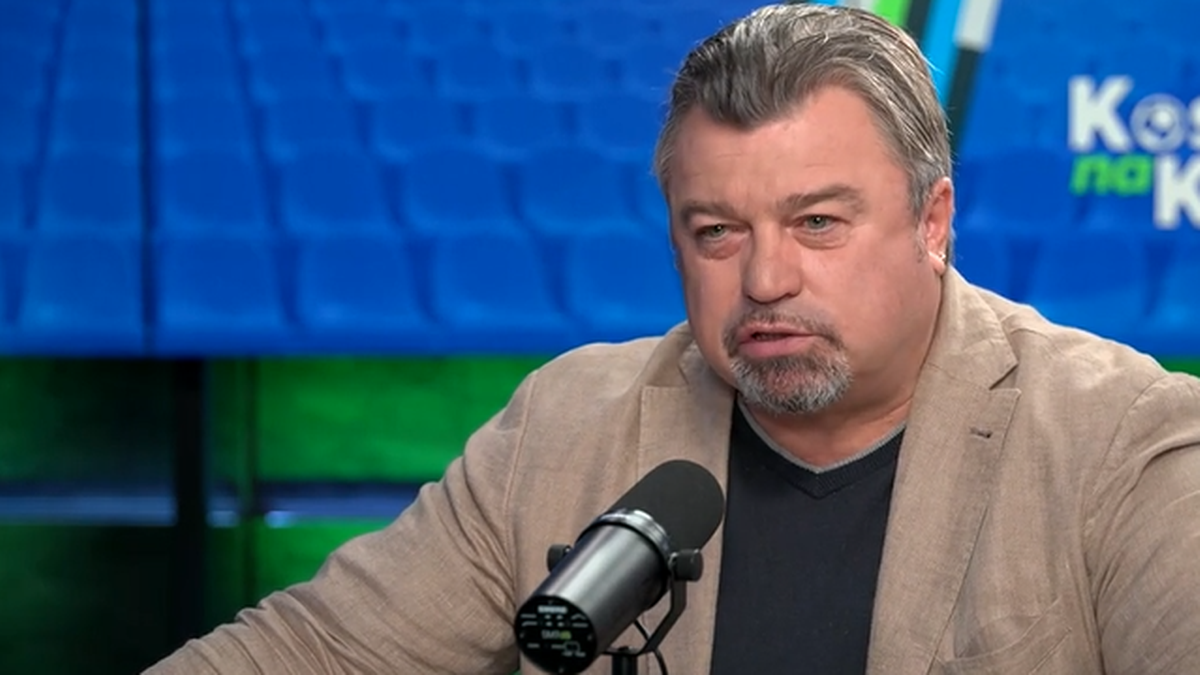 Roman Kosecki w jasnej marynarce i ciemnej koszuli, siedzi przed mikrofonem w studiu telewizyjnym. W tle widać niebieskie siedziska.