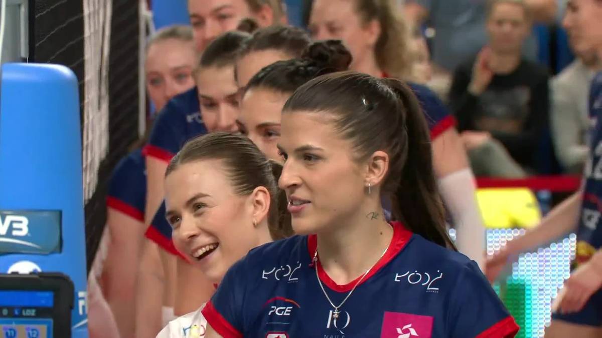 Grupa siatkarek w niebieskich strojach sportowych, z przodu dwie zawodniczki, jedna uśmiechnięta.