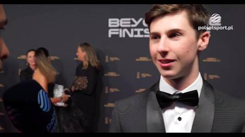 Maximo Quiles w czarnym smokingu z czarną muszką, uśmiecha się podczas wywiadu na tle czerwonego dywanu.