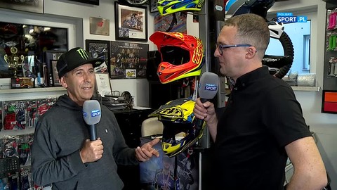 Dwóch mężczyzn rozmawia, jeden z nich trzyma mikrofon. W tle znajdują się kaski motocyklowe i akcesoria sportowe.