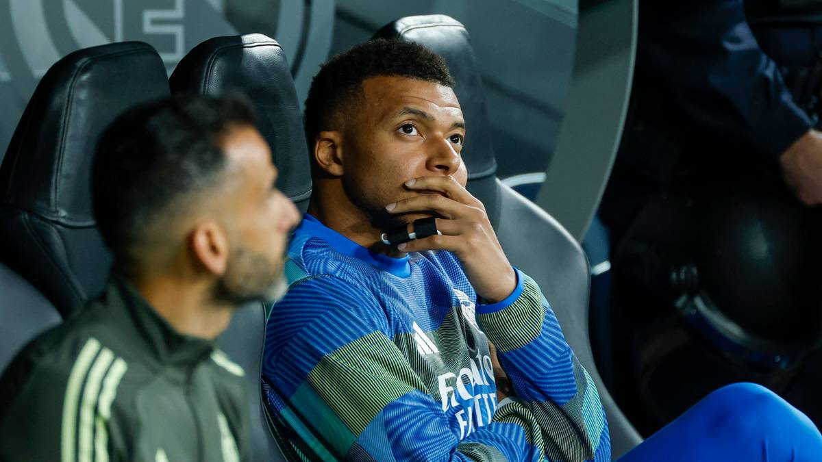 Piłkarz siedzi na ławce rezerwowych, patrząc w bok z zamyśloną miną. Ma na sobie niebiesko-białą bluzę sportową.