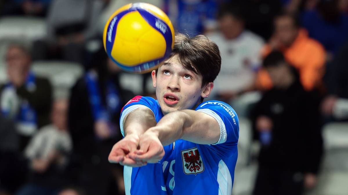 Młody siatkarz w niebieskiej koszulce z białą obwódką, w pozycji odbioru piłki, z żółto-niebieską piłką siatkową na pierwszym planie. W tle widownia.