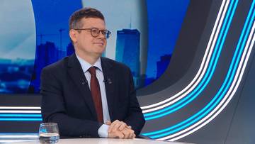 Mężczyzna w garniturze i okularach siedzi przy stole, patrząc w bok z lekko uśmiechniętą twarzą. W tle widoczny jest niebieski ekran z sylwetkami budynk&oacute;w.