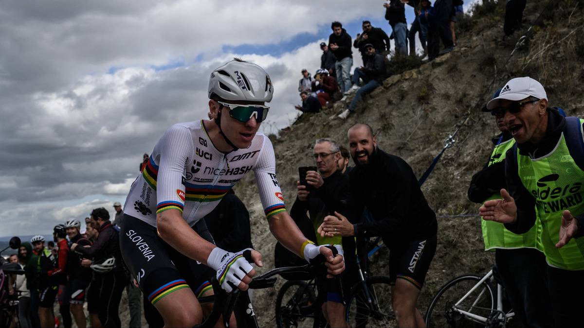 Kolarz w białej koszulce z wielokolorowymi pasami na ramionach, czapce i okularach jedzie rowerem po g&oacute;rskiej drodze. Na drugim planie widać ludzi kibicujących na zboczu g&oacute;ry.