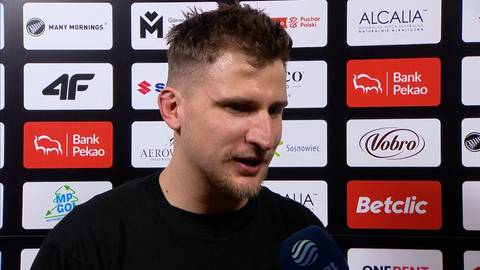 Portret Jakuba Schenka, koszykarza, z kr&oacute;tką fryzurą, m&oacute;wiącego do mikrofonu na tle ścianki sponsorskiej z logotypami.