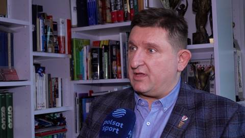 Paweł Szabelewski, mężczyzna w średnim wieku w marynarce w kratę z przypinką Polski, udziela wywiadu przed p&oacute;łkami z książkami i pucharami.