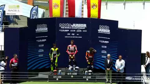 Trzech zawodnik&oacute;w na podium po zawodach motocyklowych. Zwycięzca w ż&oacute;łto-czarnym stroju na najwyższym stopniu, obok niego dwaj inni zawodnicy. Na tle flagi Hiszpanii i logo zawod&oacute;w Moto Junior FIM World Championship.