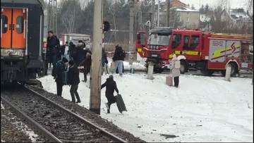Na zdjęciu widać pociąg stojący na torach kolejowych, obok kt&oacute;rego znajdują się wozy strażackie. W tle widoczni są ludzie, wśr&oacute;d nich pasażerowie z bagażami, niekt&oacute;rzy w towarzystwie służb ratowniczych.