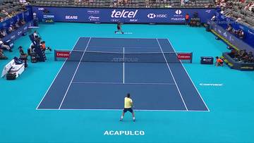 Dwaj tenisiści na niebieskim korcie podczas meczu. Jeden zawodnik w żółtej koszulce na pierwszym planie, drugi po drugiej stronie siatki. W tle trybuny z widzami i banery sponsorów.