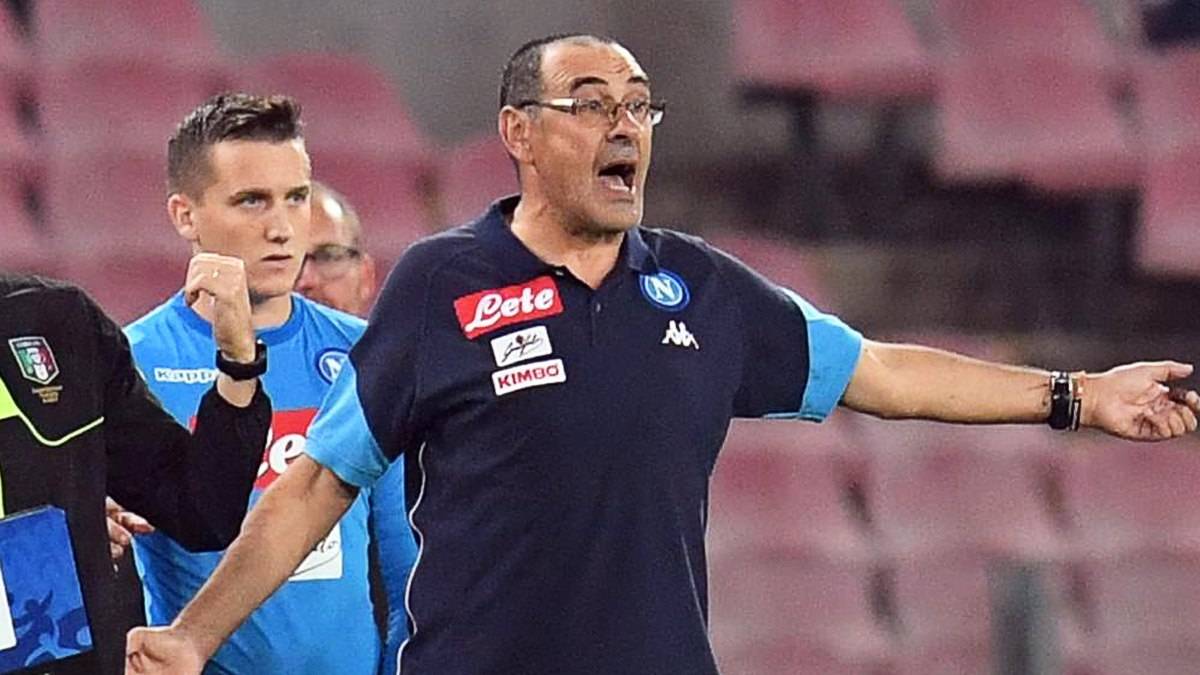 Trener piłkarski w okularach i granatowej koszulce z logo Napoli gestykuluje na tle stadionu.